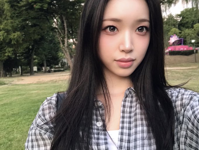 A selfie-style portrait of a young woman with long dark hair, wearing a black and white plaid shirt over a white top, outdoors in a park setting.