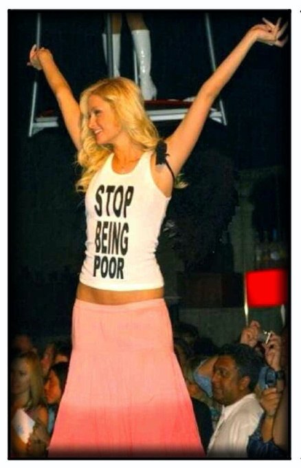 A blonde woman stands on a stage with her arms raised, wearing a white tank top that reads 'STOP BEING POOR' and a long pink skirt.