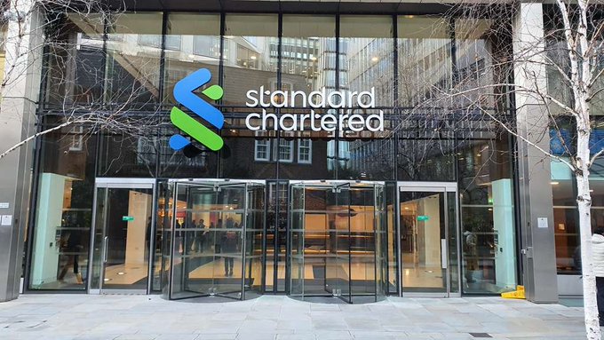 The image shows a modern glass building, likely the entrance of a bank or financial institution like Standard Chartered, featuring large windows that reflect outdoor surroundings. The sign above the glass doors prominently displays "STANDARD CHARTERED" followed by "BITCOIN BOTTOMED AT $59K". This indicates that there is a Bitcoin trading opportunity available within the premises of the building. The focus on both the exterior appearance and the offerings inside demonstrates the financial services offered at this establishment.