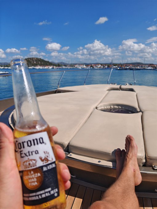 A hand holds a Corona Extra beer bottle while relaxing on a boat, with bare feet visible and a coastal town in the background under a blue sky.