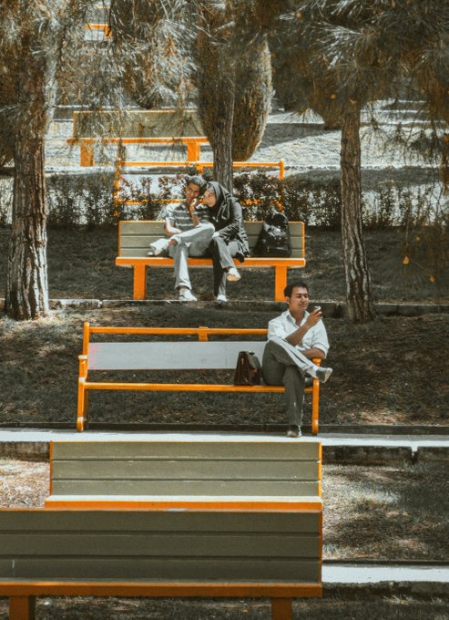 A park scene with desaturated tones and orange-tinted benches shows a couple sitting together on one bench while a man in a white shirt looks at his phone on another bench in the foreground.