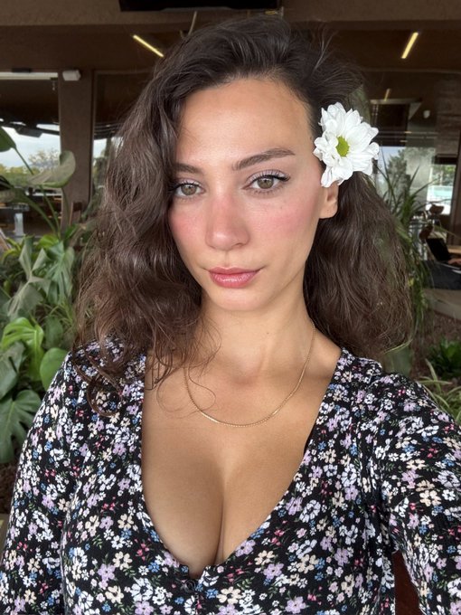 A close-up portrait of a young woman with wavy brown hair, a white flower tucked behind her ear, and a floral-patterned top.