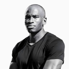 A black-and-white portrait of a muscular, bald African American man wearing a tight black V-neck t-shirt, looking directly at the camera.