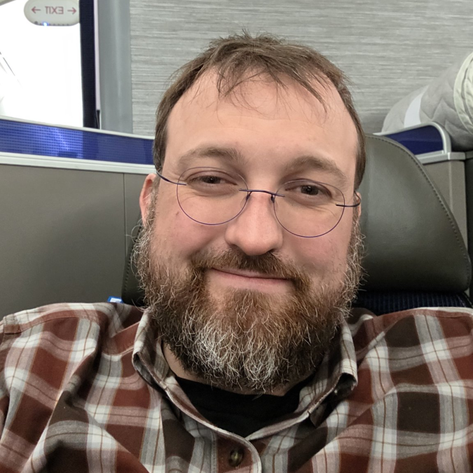 The image features a man with glasses, a beard, and a plaid shirt sitting comfortably in an airplane seat. He appears to be smiling at the camera, possibly posing for a photo or video. The background of the image shows a window, which could indicate that they are on a flight. There is also text visible in the image, which reads "good news for Cardano founder he still has two years". This suggests that the man might have some connection to the Cardano cryptocurrency or blockchain project and is excited about its future.