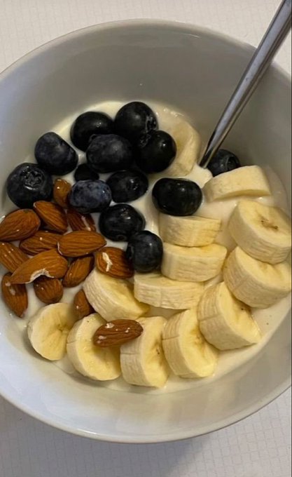 A bowl of cereal with milk, topped with sliced bananas, blueberries, and almonds, with a spoon resting inside.