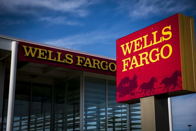 A Wells Fargo bank branch sign displays the company's red and yellow logo featuring a horse-drawn stagecoach. The image relates to Wells Fargo's new initiative to offer tokenized deposits, representing its move toward digital banking solutions for corporate clients.