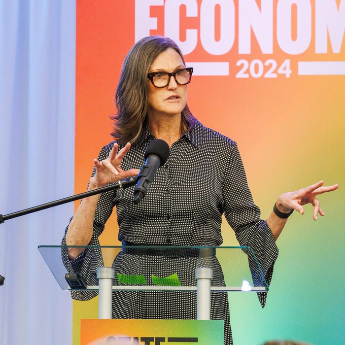 Cathie Wood, CEO of ARK Invest, speaks at a podium during an event with "ECONOMY 2024" displayed on the colorful backdrop behind her, gesturing with her hands as she makes her case for Bitcoin as a transformative technology and monetary system.