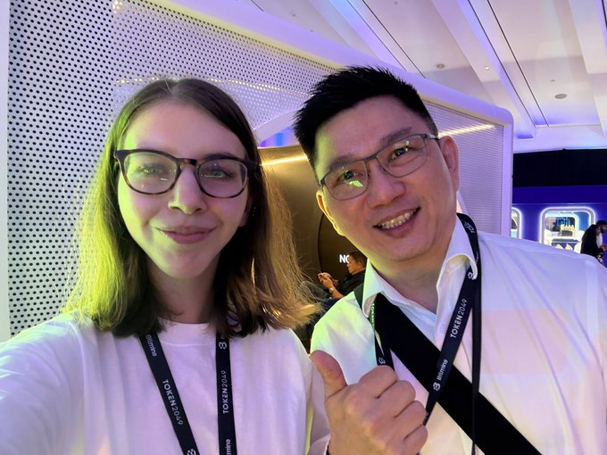 A woman with glasses and an East Asian man wearing glasses pose together at an event, both wearing lanyards with 'TOKEN2049' branding.