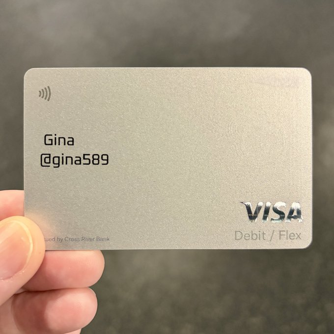 A hand holds a minimalist silver Visa Debit / Flex card bearing the name 'Gina' and the handle '@gina589', issued by Cross River Bank.