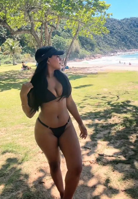 A woman in a black bikini and baseball cap stands on a grassy area near a tropical beach, smiling as she looks to the side. The caption "Happy Sunday, Let's go touch some grass?" playfully ties into the lush green grass beneath her feet in the sunny coastal setting.