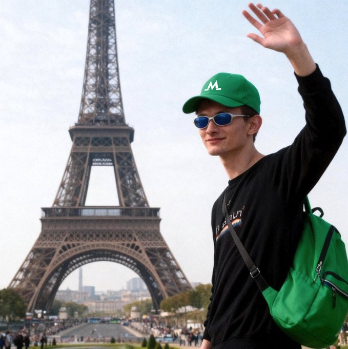 A man wearing a green "M" cap, blue sunglasses, and a black shirt waves in front of the Eiffel Tower in Paris, with a green bag slung over his shoulder.