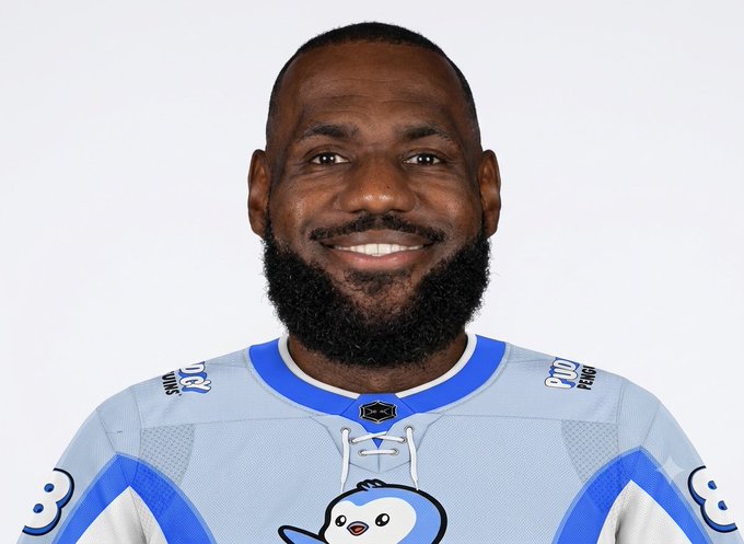 A headshot of LeBron James smiling, wearing a light blue and white jersey featuring a penguin/cartoon character logo.