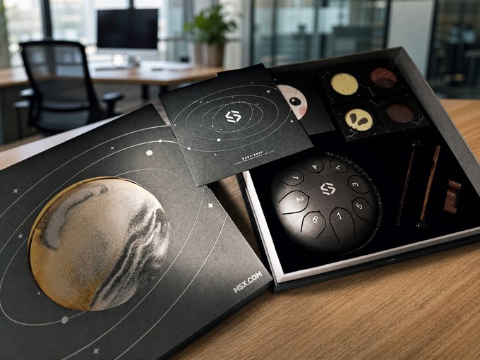 An open MSX-branded product box on a wooden desk, featuring a black spherical device with numbered buttons, alongside a chocolate assortment tray and a card depicting the planet Saturn.