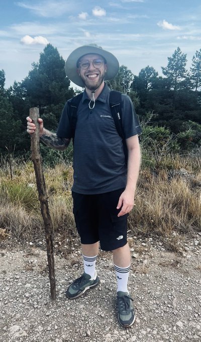 A bearded hiker wearing a Columbia shirt, New Balance shorts, and long striped socks stands smiling on a gravel trail while holding a wooden walking stick, looking out of place in the wilderness. The caption humorously suggests that this unassuming outdoorsman represents the easy competition you'd face in trading.