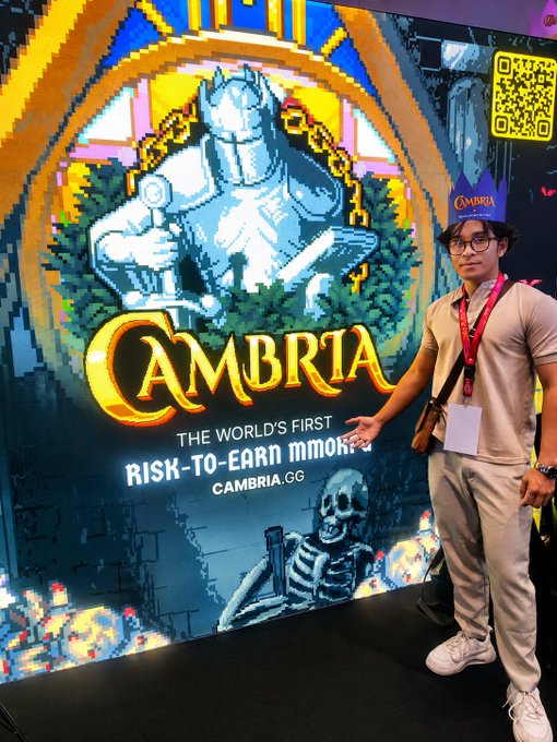 A person wearing glasses and a lanyard poses next to a colorful pixel-art promotional banner for "CAMBRIA," described as "THE WORLD'S FIRST RISK-TO-EARN MMO" with the website "CAMBRIA.CG" displayed, reflecting their Web3 gaming and community-building background.