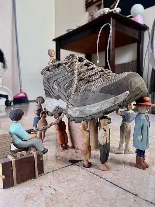 A real-life photograph shows a large, muddy hiking boot tied with a rope and surrounded by several small, carved wooden figurines of people, appearing to lift or support the shoe on a tiled floor.