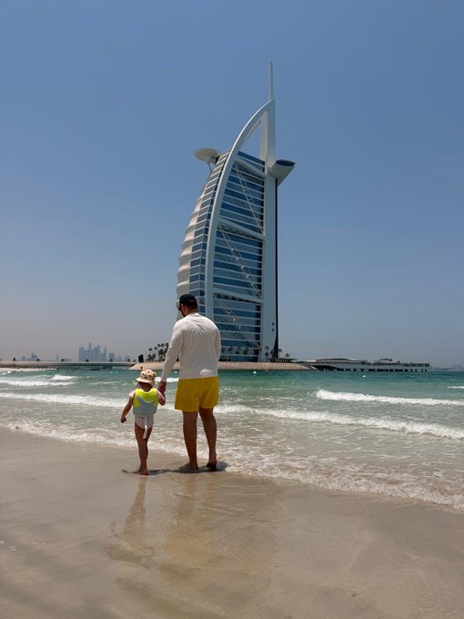 A father and child stand hand-in-hand on Dubai's beach, with the iconic Burj Al Arab towering in the background as they gaze out at the ocean. The serene family moment complements the post's warm message, emphasizing the value of family and friends shared alongside a sunny Dubai greeting.