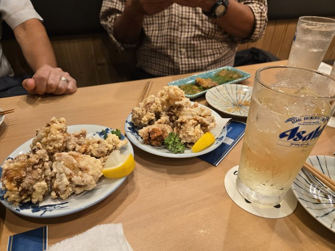 A dining scene with two plates of karaage (fried chicken) garnished with lemon, glasses of Asahi beer, and other dishes on a wooden table.