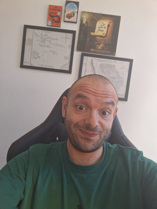 A bald man with a beard wearing a green t-shirt sits in a gaming chair, with framed drawings, maps, and books including 'Le Petit Nicolas' and 'De Sable & de Braise' hanging on the wall behind him.