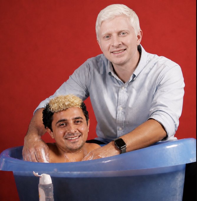 A man with blond hair soaks in a blue tub while another man, also with blond hair, leans over him and places his hands on his shoulders. The pairing of these two individuals in a domestic setting, perhaps implying a fresh start or a cleansing, likely relates to the post's claim that they will "bring SOL back."