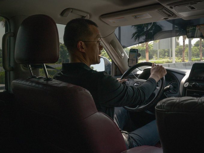 A man in a dark long-sleeved shirt and glasses drives a car with burgundy leather seats, glancing over at a smartphone mounted on the dashboard displaying navigation. The image suggests CZ, founder of Binance, en route to the TOKEN2049 crypto conference, with the sunny, palm-tree-lined street visible through the windows hinting at travel to a major industry event.