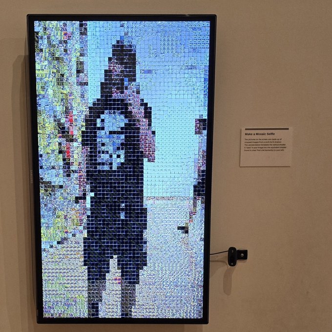 A digital mosaic artwork titled "Make a Mosaic Selfie" displays a pixelated image of a child holding a teddy bear, displayed on a screen next to its description placard.