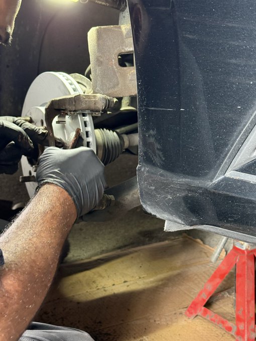 A mechanic wearing black gloves works on a vehicle's brake assembly while the car is supported by a red jack stand in a garage setting.