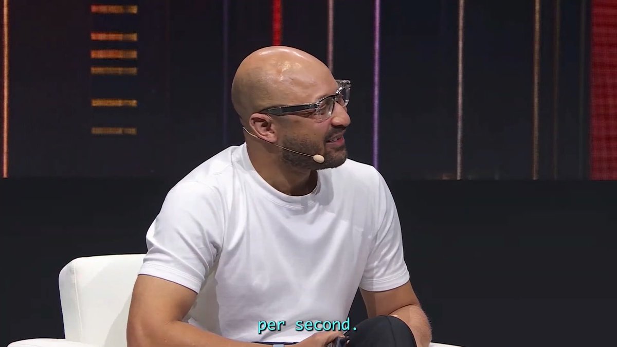 A bald man with glasses and a beard, wearing a white t-shirt and headset microphone, speaking on a stage set with vertical light bars; the subtitle reads 'per second.'