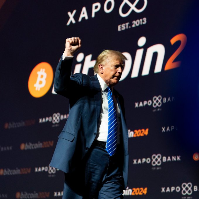 A photograph of Donald Trump raising his fist at what appears to be a Bitcoin 2024 conference, with XAPO and Bitcoin logos visible on the backdrop behind him.
