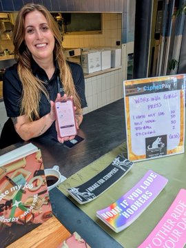 A smiling woman with long hair holds up a smartphone at a coffee shop counter, with a sign reading 'Crispy Pies' advertising 'Vegan Thai Fries' visible alongside promotional stickers for 'HOPE IF YOU LOVE HOPPERS' and other food items.