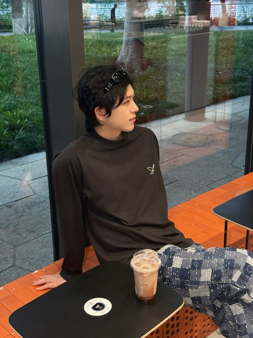 A young man with sunglasses on his head sits at an outdoor cafe table with an iced coffee, wearing a black sweatshirt and patterned pants.