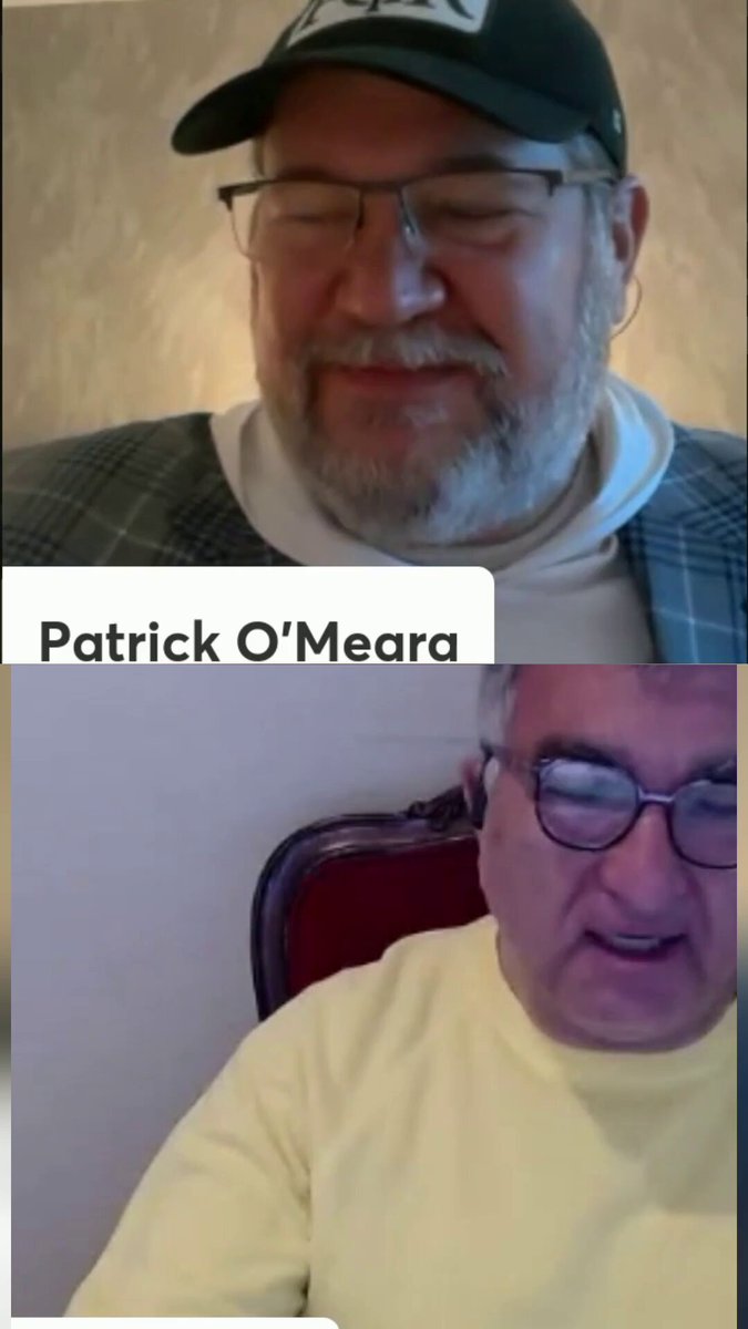 The image shows two men sitting next to each other, both wearing glasses and a hat. They are engaged in an online video call with the names "Patrick O'Meera" and "Jean Paoli" displayed at the top of the screen. The background features a wall painted white, providing a neutral backdrop for their conversation.