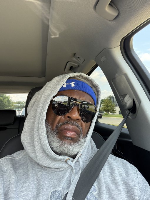 A man with a gray beard wearing a gray hoodie, blue Under Armour headband, and aviator sunglasses, sitting in the driver's seat of a car with his seatbelt on.