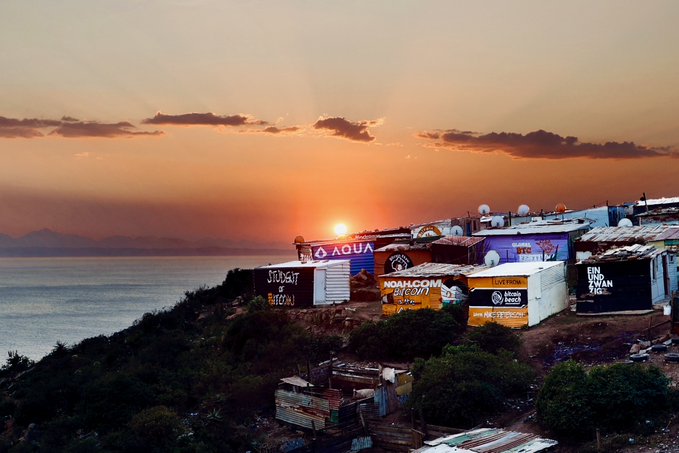 A scenic sunset view over the ocean with buildings on a cliffside displaying advertisements including 'AQUA' and 'NOAH.COM OM 2024'.