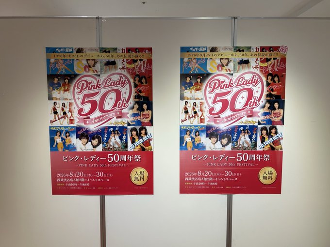 Two identical promotional posters for the "Pink Lady 50th Anniversary" exhibition (ピンク・レディー 50周年祭) hang on a white wall, featuring the duo's iconic pink heart logo and photos spanning from their 1976 debut through their 1980s comeback, with event details indicating an August 20-30, 2026 run at Tokyu Plaza Akasaka (東急百貨店A館2階). The posters relate to the poster's nostalgic memory of seeing the Pink Lady 50th anniversary exhibition from their childhood.