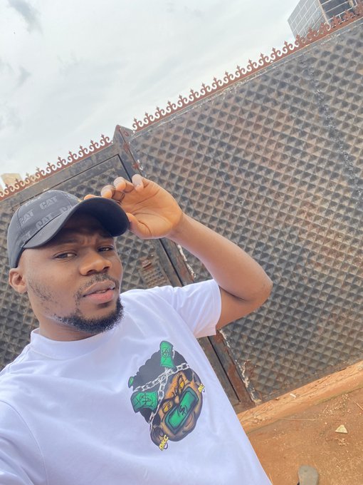 A selfie of a man wearing a white graphic t-shirt and a black cap standing in front of a large metal gate with decorative spikes.