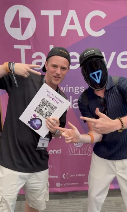 Two people posing at a 'TAC' event, one wearing a black t-shirt and cap holding a QR code flyer to 'Scan to connect with TAC', and another wearing a black helmet with a blue triangle logo.
