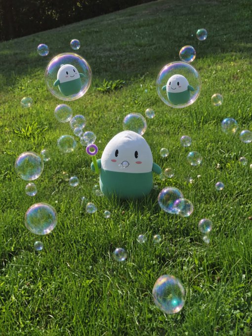 A 3D-rendered scene of a cute round mascot character with a white face and green body blowing bubbles on a grassy lawn, with smaller versions floating inside the bubbles.