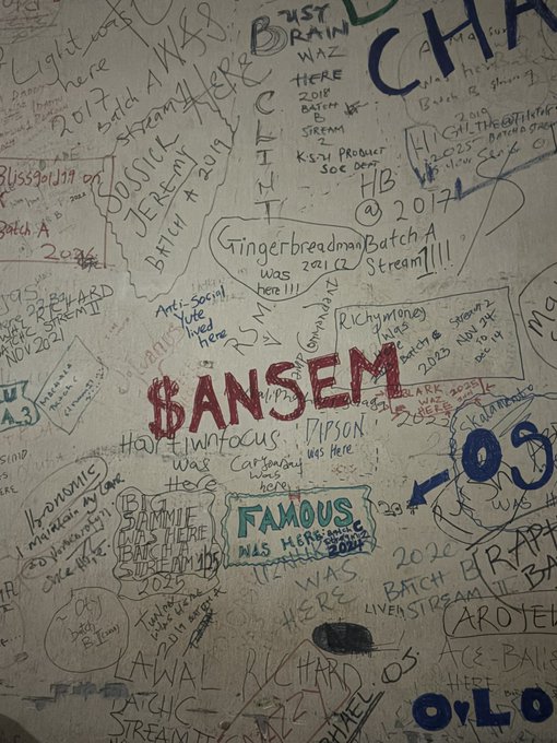 A wall covered in graffiti and marker signatures from various internet personalities, prominently featuring the name 'ANSEM' in red, along with 'FAMOUS' and references to 'Batch A' streams from 2022-2023.
