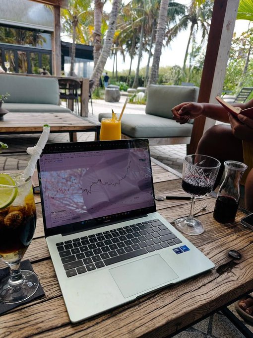 A laptop displaying a stock market chart sits on a wooden table at an outdoor cafe, surrounded by glasses of dark and orange beverages with palm trees in the background.