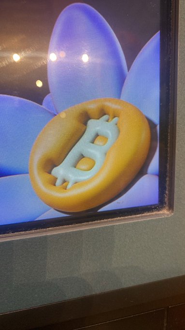 A digital display shows a 3D rendering of a Bitcoin (₿) symbol, depicted in light blue and embossed on a golden coin-shaped object, set against a backdrop of large blue flower petals.