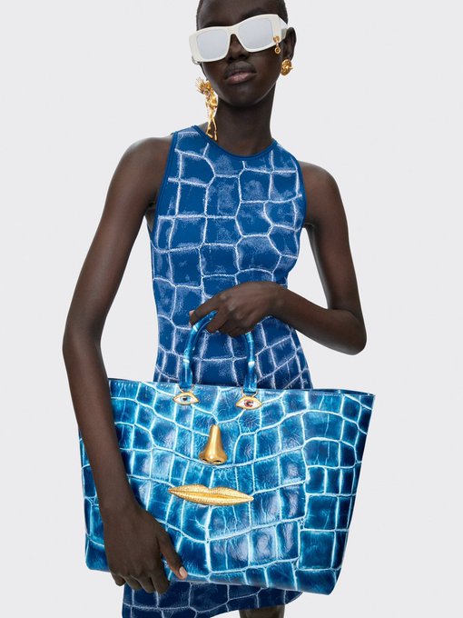 A model poses wearing white visor sunglasses, gold statement earrings, and a blue crocodile-embossed sleeveless dress, while holding a matching blue crocodile-patterned tote bag featuring whimsical face details with gold accents.