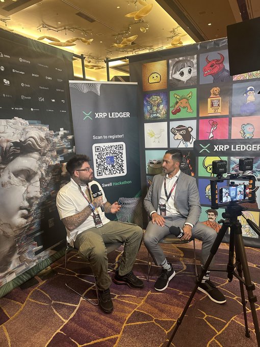 Two men are seated for an interview in front of an XRP Ledger banner featuring a QR code and NFT artwork, with a camera on a tripod filming them.
