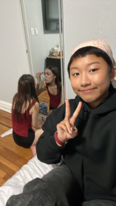 A selfie of a smiling person wearing a head covering, while another person sits in front of a mirror applying makeup in the background.