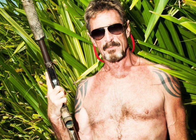 A shirtless man with sunglasses, a mustache, tribal tattoos, and an earpiece stands holding a rifle in front of tropical palm foliage.