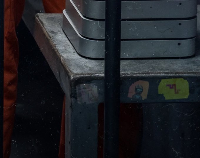 A close-up view shows a worn metal table or equipment with stickers, including one resembling the Statue of Liberty, set against a dark background with red fabric.