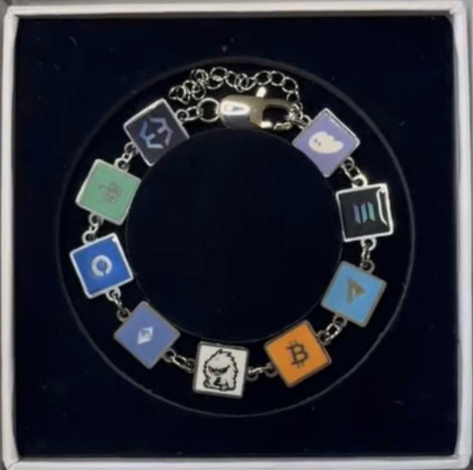 A silver charm bracelet displayed in a black gift box features square pendants representing various platforms and cryptocurrencies, including Bitcoin (₿), along with recognizable app icons. The post "I need one" suggests the user is expressing a desire to own this tech/crypto-themed accessory.
