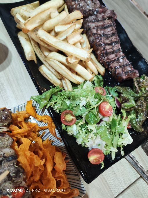A meal featuring grilled steak, French fries, a fresh green salad with cherry tomatoes, and sautéed green peppers, with additional meat and crispy sweet potato chips on the side; watermarked 'XIAOMI 12T PRO | @SACRA97'.