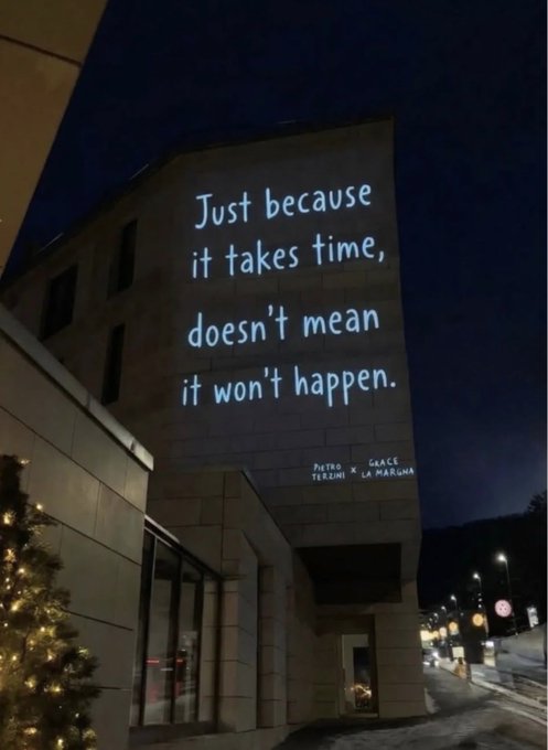 A nighttime photograph of a building facade features a glowing projected text that reads, 'Just because it takes time, doesn't mean it won't happen,' with a Christmas tree visible in the lower left.