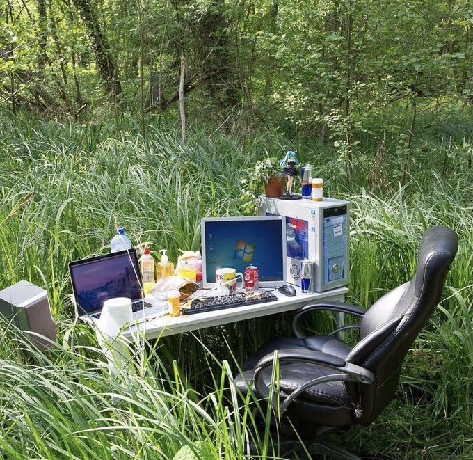 A makeshift outdoor computer desk setup in a grassy field, complete with a laptop, monitor displaying Windows, keyboard, tower PC, office chair, and various snacks and drinks.